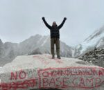 Person standing on a stone at Everest Base Camp with snow-capped Himalayan peaks in the background under a clear blue sky.