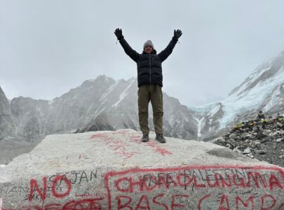Person standing on a stone at Everest Base Camp with snow-capped Himalayan peaks in the background under a clear blue sky.