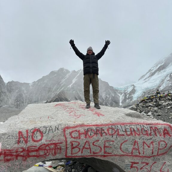 Person standing on a stone at Everest Base Camp with snow-capped Himalayan peaks in the background under a clear blue sky.
